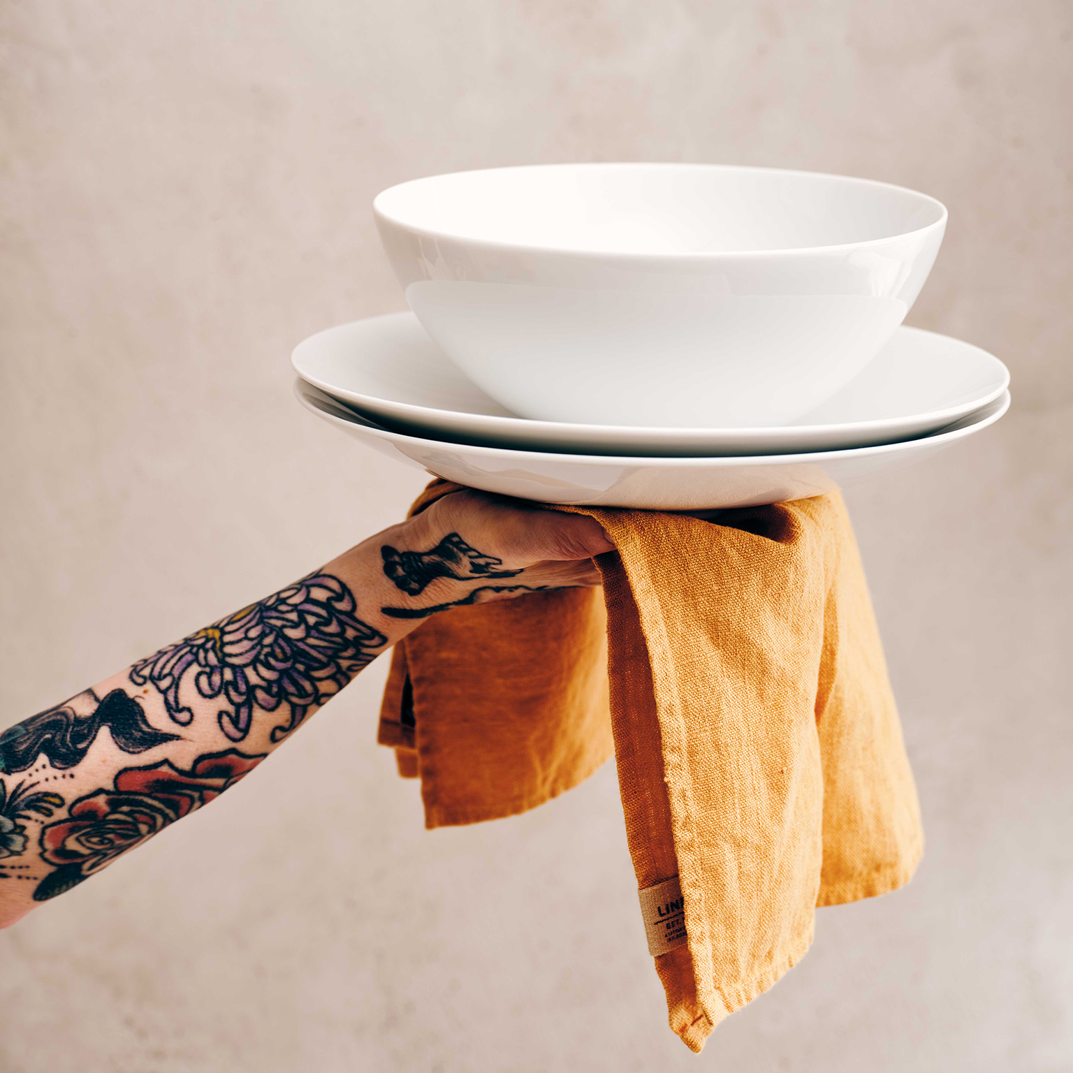 A tattooed arm on a napkin holds the TAC Allround bowl stacked on TAC Allround plate in the air.
